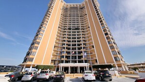 A high-rise hotel with multiple balconies, a flag, and a parking area with cars. - Wake up to beautiful ocean views!!! (MYRTLE BEACH)