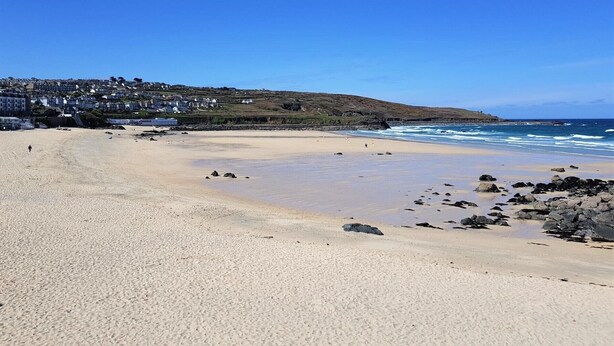 A sandy beach with scattered rocks, a clear blue sky, and a hillside with buildings in the background.