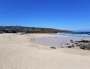 Una playa arenosa con rocas dispersas, un cielo azul despejado y una colina con edificios al fondo.