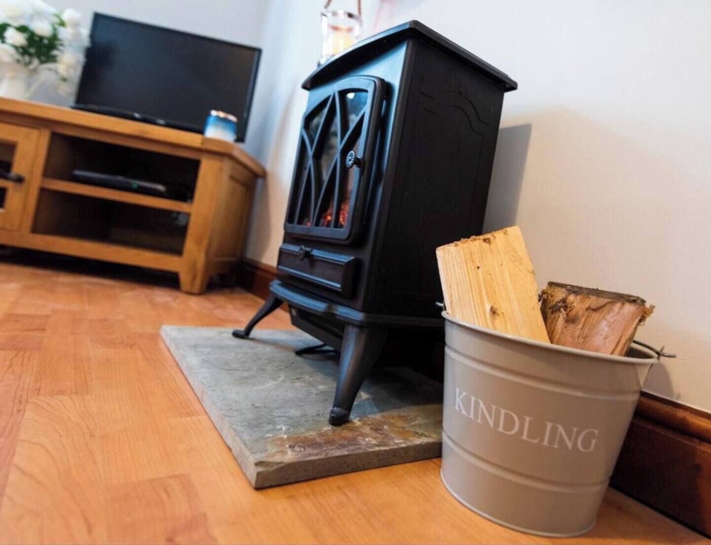 A black wood stove with a metal bucket labeled "KINDLING" beside it, placed on a wooden floor in a room with a wooden cabinet and a television in the background.