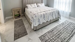 Un dormitorio con cama, ventilador de techo, mesita de noche, un cuadro enmarcado y una ventana con cortinas.