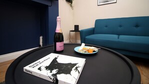 A living room with a blue sofa, a black coffee table, a book titled 'Banksy V'ill and Pieces', a bottle of champagne, and a plate with cheese and crackers. - Karah Suites - Church Road I (Bristol)