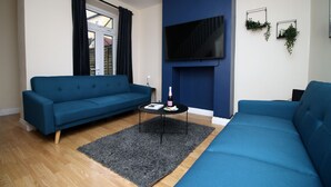 A modern living room with two blue sofas, a flat-screen TV mounted on the wall, and a small table with a bottle and a book. - Karah Suites - Church Road I (Bristol)