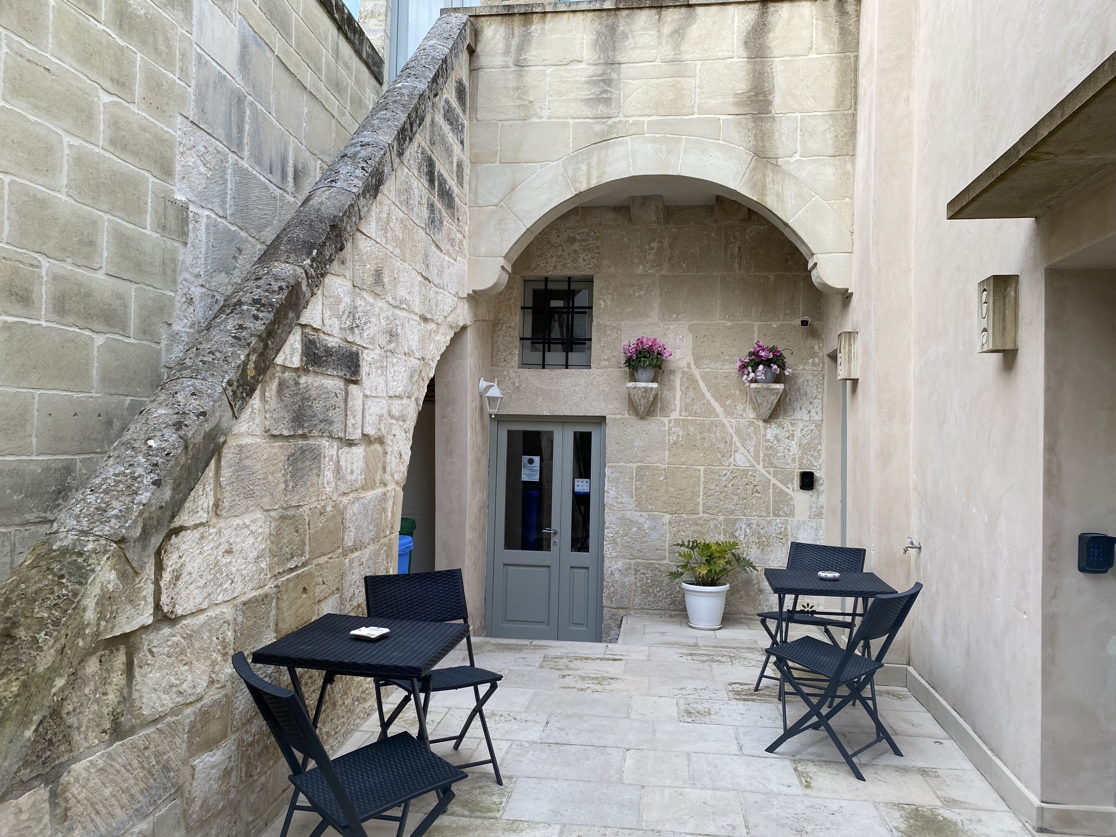 Um edifício de pedra com entrada em arco, uma pequena varanda com mesa e cadeiras, e plantas em vasos.