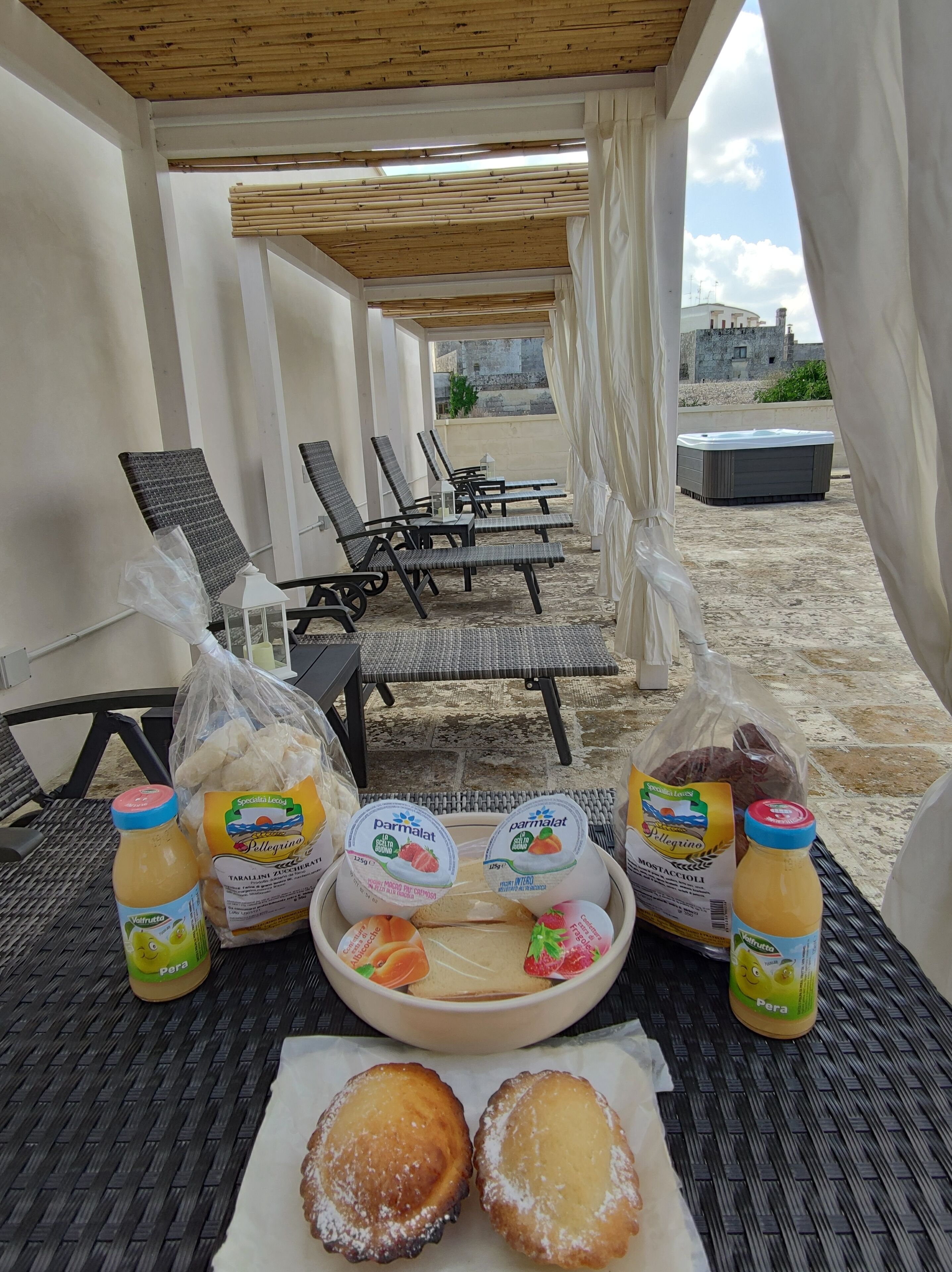 Un espace de détente aménagé sur la plage, avec des fauteuils en osier, une table garnie de nourriture et de boissons, et une vue sur la mer et les bâtiments en arrière-plan.