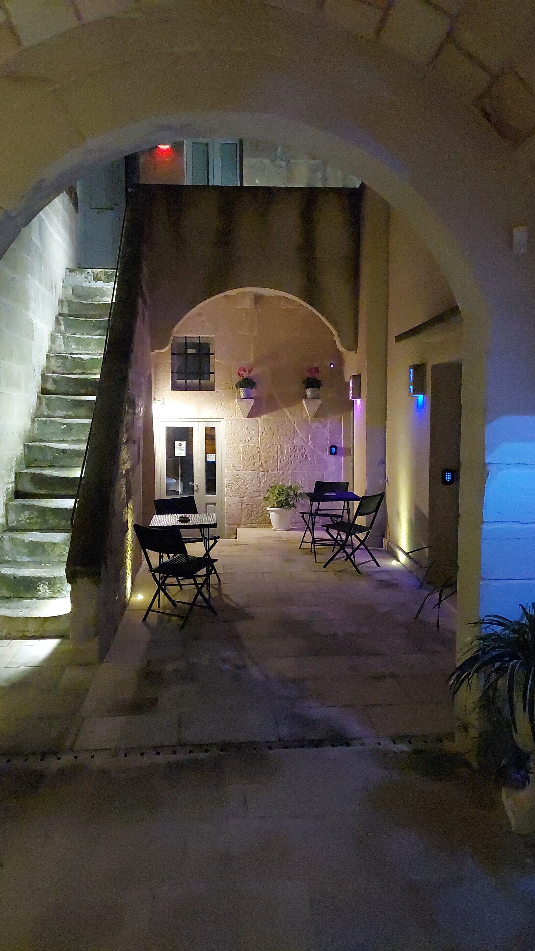 Un escalier en pierre mène à une pièce où se trouvent un lit, une petite table et deux chaises. On y trouve également des plantes en pot et un luminaire fixé au mur.