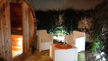 Eine Holzsauna mit weißem Innenraum, ein weißer Außenbereich mit Tisch und eine überdachte Terrasse mit Grünpflanzen.