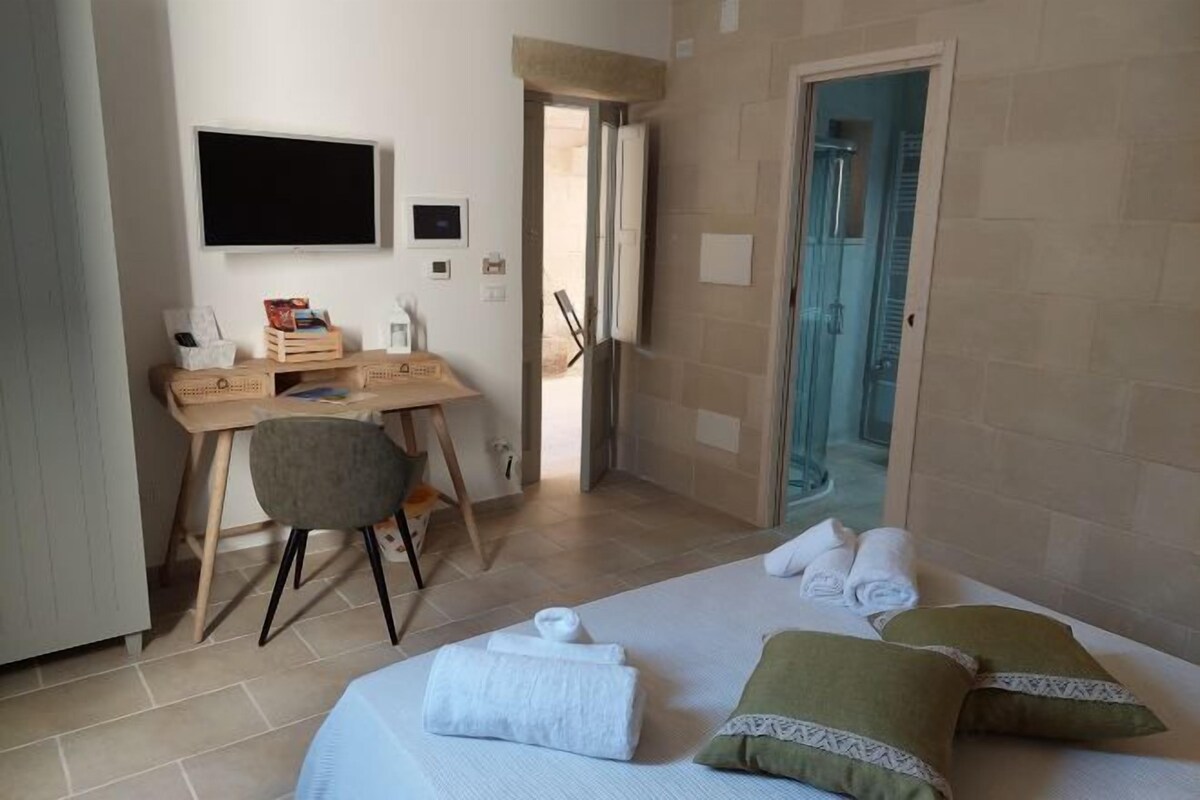 A hotel room with a bed, a desk, a chair, a TV, and a bathroom visible through an open door.