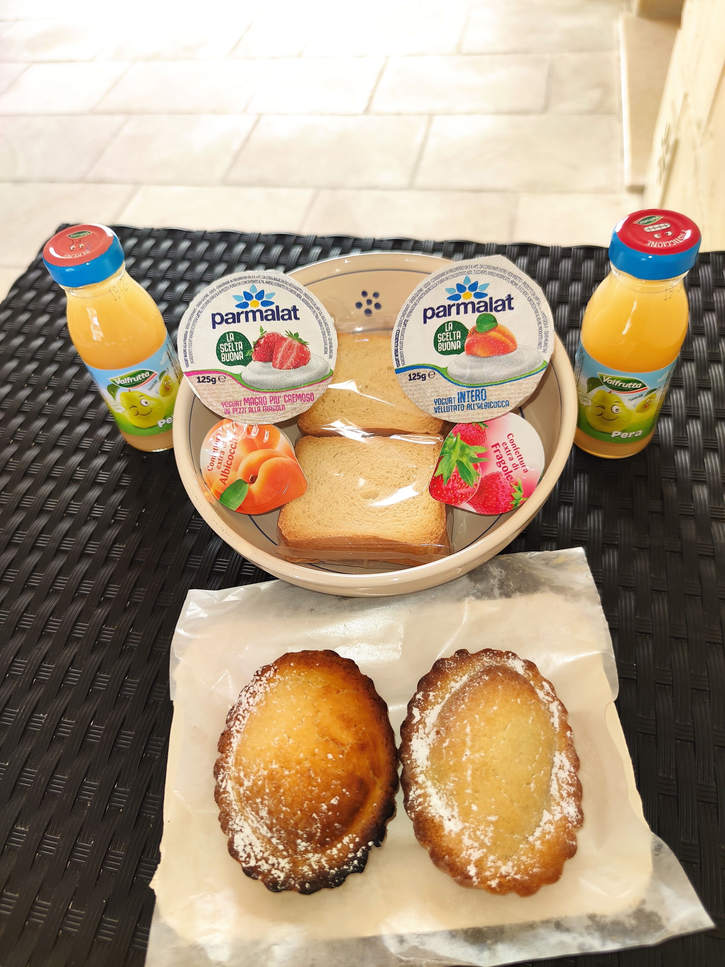 A breakfast tray with two bottles of orange juice, two yogurts, a slice of bread, a strawberry, and two pastries on a woven table.