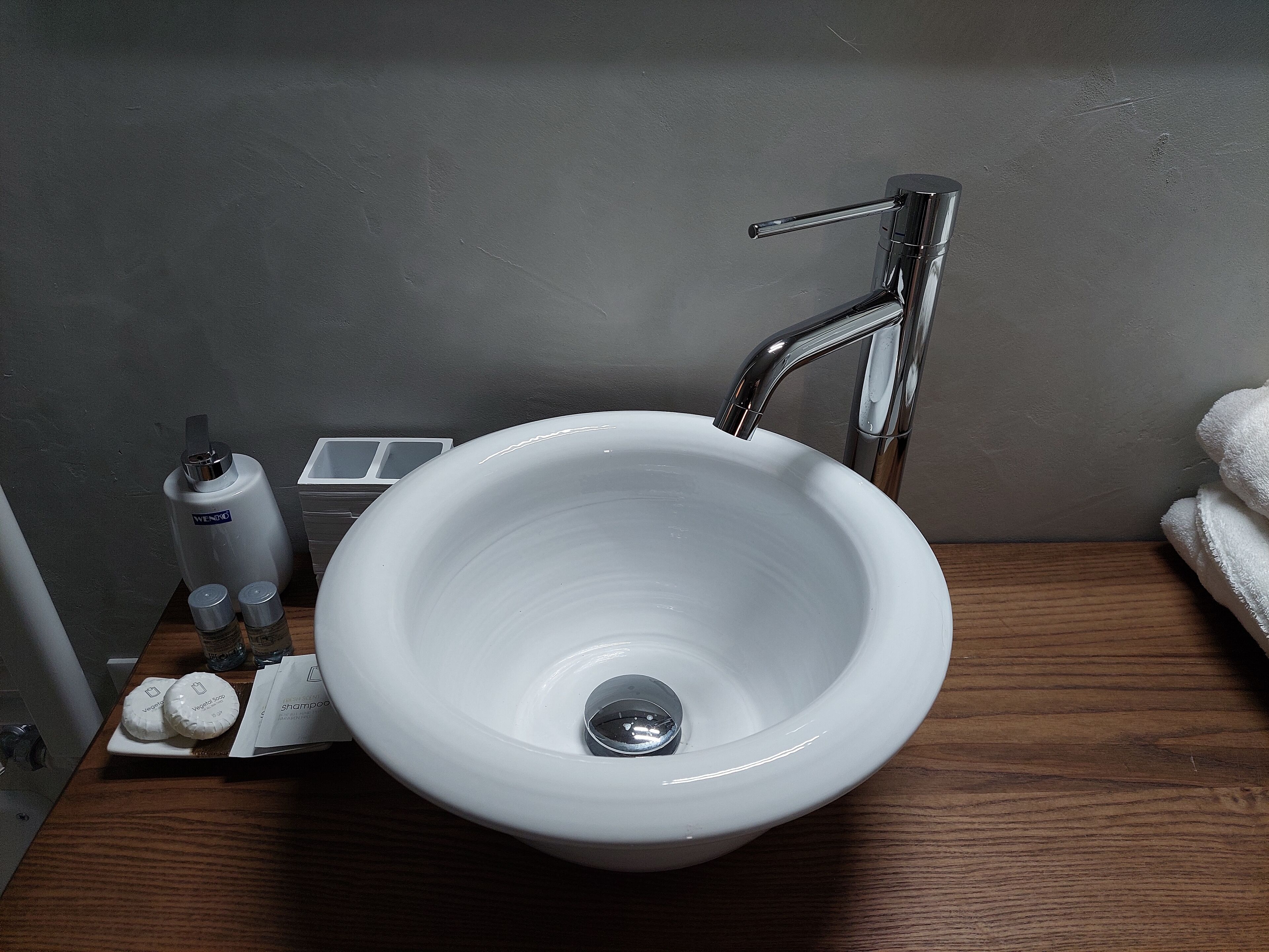 A white ceramic sink with a chrome faucet, a soap dispenser, and a bottle of shampoo on a wooden countertop.