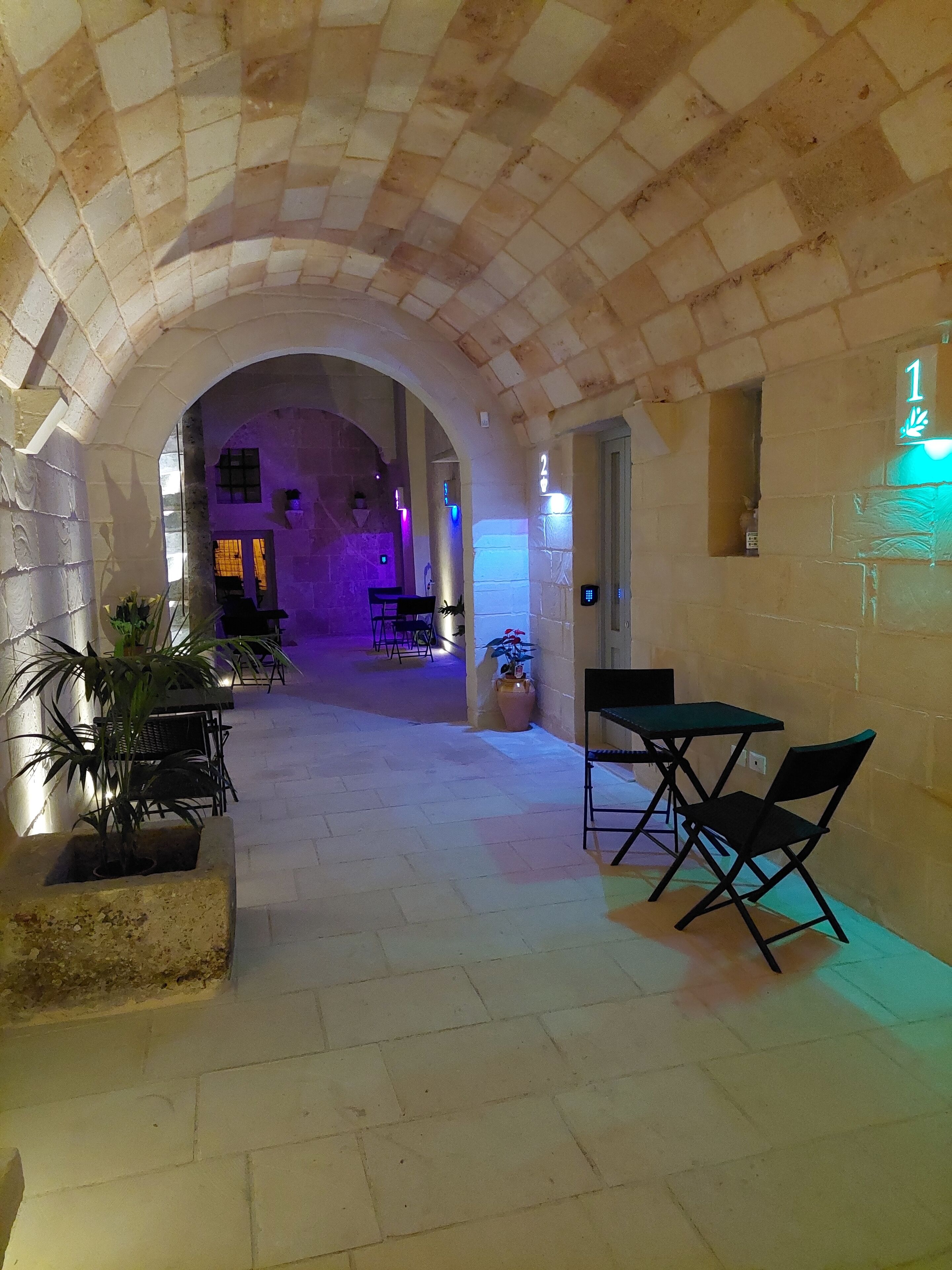 An arched corridor with stone walls, a seating area with tables and chairs, and a potted plant.