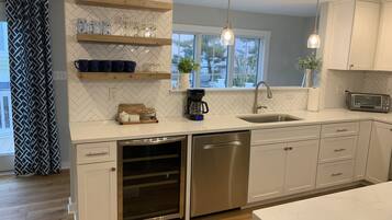 A modern kitchen with white cabinetry, a dishwasher, a built-in oven, and a microwave. There are wooden shelves with glassware and a coffee maker.