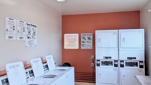 Un espace buanderie équipé de plusieurs machines à laver et de plusieurs sèche-linge, ainsi que de panneaux d’information sur les services proposés et d’un simple panneau indiquant l’endroit où étendre le linge.