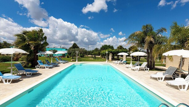 A swimming pool with clear blue water, surrounded by white sun loungers and umbrellas, with palm trees and a clear sky in the background. - Tenuta Donna Caterina by BarbarHouse (Mesagne)