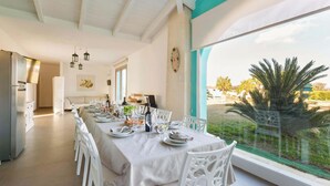 A dining area with a long table set for a meal, white chairs, and a view of the outdoors through large windows. - Tenuta Donna Caterina by BarbarHouse (Mesagne)