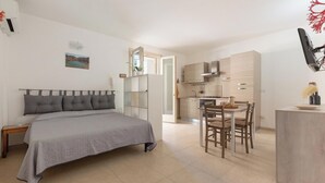Une chambre moderne avec un grand lit, une petite table de chevet et une commode. On y trouve également un coin cuisine avec une table et des chaises, ainsi qu’un petit îlot central doté d’un évier.