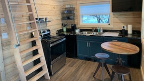 A modern kitchen with wooden paneling, a stove, a sink, and a window overlooking a snowy landscape. - Downtown Cozy Red Cabin (Victor)