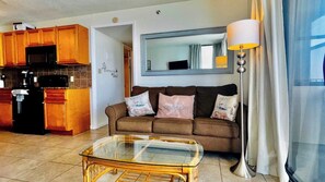 A living room with a glass coffee table, a sofa with cushions, a lamp, and a kitchen area with wooden cabinets and appliances.