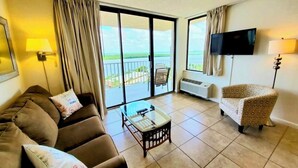 A living room with a sofa, armchair, and coffee table, large windows with curtains, a flat-screen TV, and a view of the outdoors. - Lovers Key Beach Club 1105 - Private Beach (Bonita Springs)