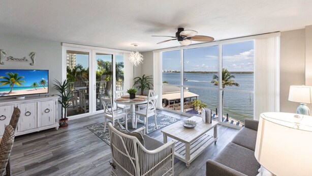 A spacious living area with a large window overlooking a body of water, a ceiling fan, and a television on a white cabinet.