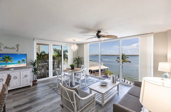 A spacious living room with a large window overlooking a body of water, a ceiling fan, and a television on a cabinet.