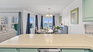 A modern kitchen with a central island, dining table, and chairs. There are large windows with curtains, a view of buildings, and a hanging light fixture.