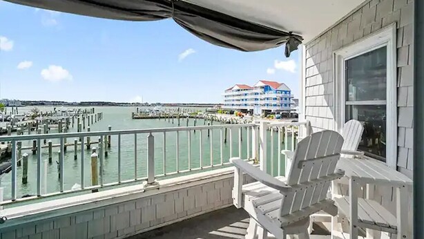 A balcony with white Adirondack chairs, a view of a marina, and a large building with a red roof in the background.