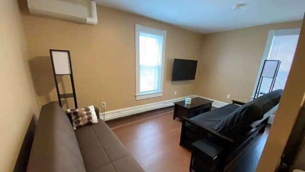 A modern living room with a sofa, a small table, a flat-screen TV mounted on the wall, and a window providing natural light. - Apt 3A Lake Bomoseen Inn “The Dog” with boat slip. (Castleton)