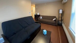 A living room with a dark sofa, a coffee table, a floor lamp, and a window with blinds. - Apt 3A Lake Bomoseen Inn “The Dog” with boat slip. (Castleton)
