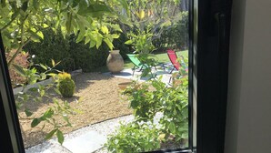A view through a window of a garden area with a gravel path, a large planter, and outdoor furniture. - Cosy studio for 2 near the beach (Sanary-sur-Mer)