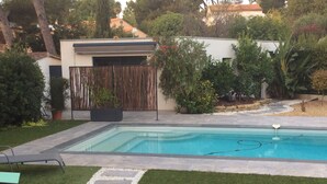 A swimming pool with a stone pathway leading to a lounge area, surrounded by lush greenery and a modern house. - Cosy studio for 2 near the beach (Sanary-sur-Mer)
