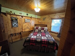 Un dormitorio de cabaña acogedor con techo de madera, una cama con colcha escocesa, una ventana con persianas y un calentador fijado a la pared.