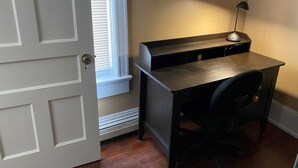 A compact office space with a desk, chair, and a lamp. A door is visible on the left, and a window with blinds is on the right. - Apt 3C Lake Bomoseen Inn “The Dog” with boat slip.  (Castleton)