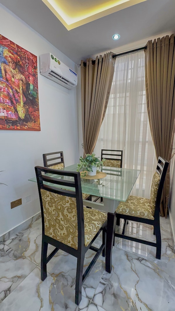 A dining area with a glass table, four chairs with patterned upholstery, a potted plant, and a colorful abstract painting on the wall. - 5 Bedrooms Einsuite House in Lekki Lagos Nigeria (Lekki)