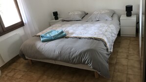 Een net opgemaakt bed met een opgevouwen blauwe handdoek, een wit beddegoed met bloemenprint en passende kussens. Twee nachtkastjes met zwarte speakers, een witte nachtkastje met een lamp, en een raam met gordijnen.