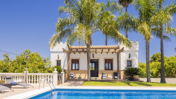 Vista de una piscina junto a un edificio con pérgola, palmeras y agua cristalina.