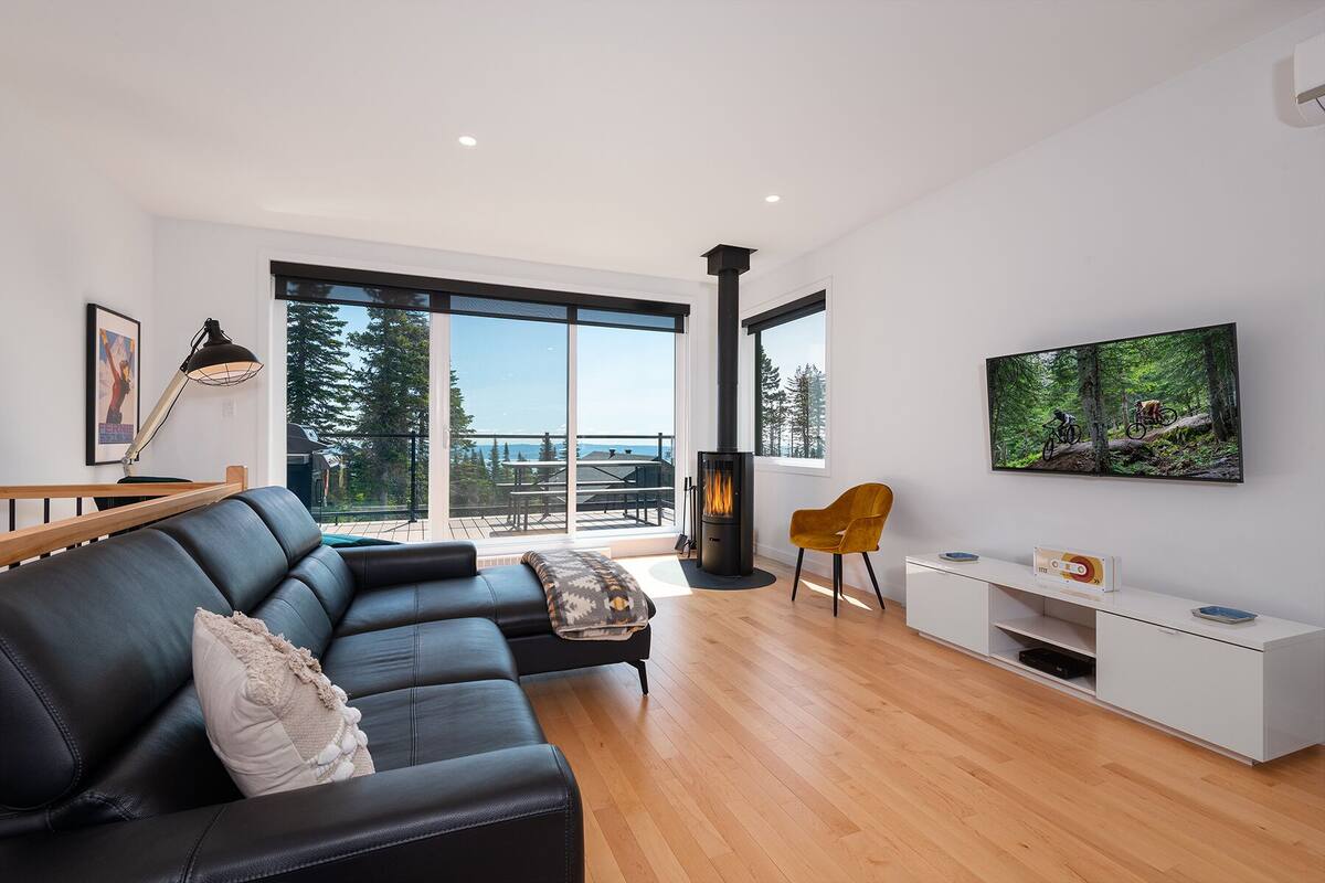 A modern living room with a black leather sofa, a wooden floor, a fireplace, a dining area with a table and chairs, and a large window offering a view of trees and a distant landscape.