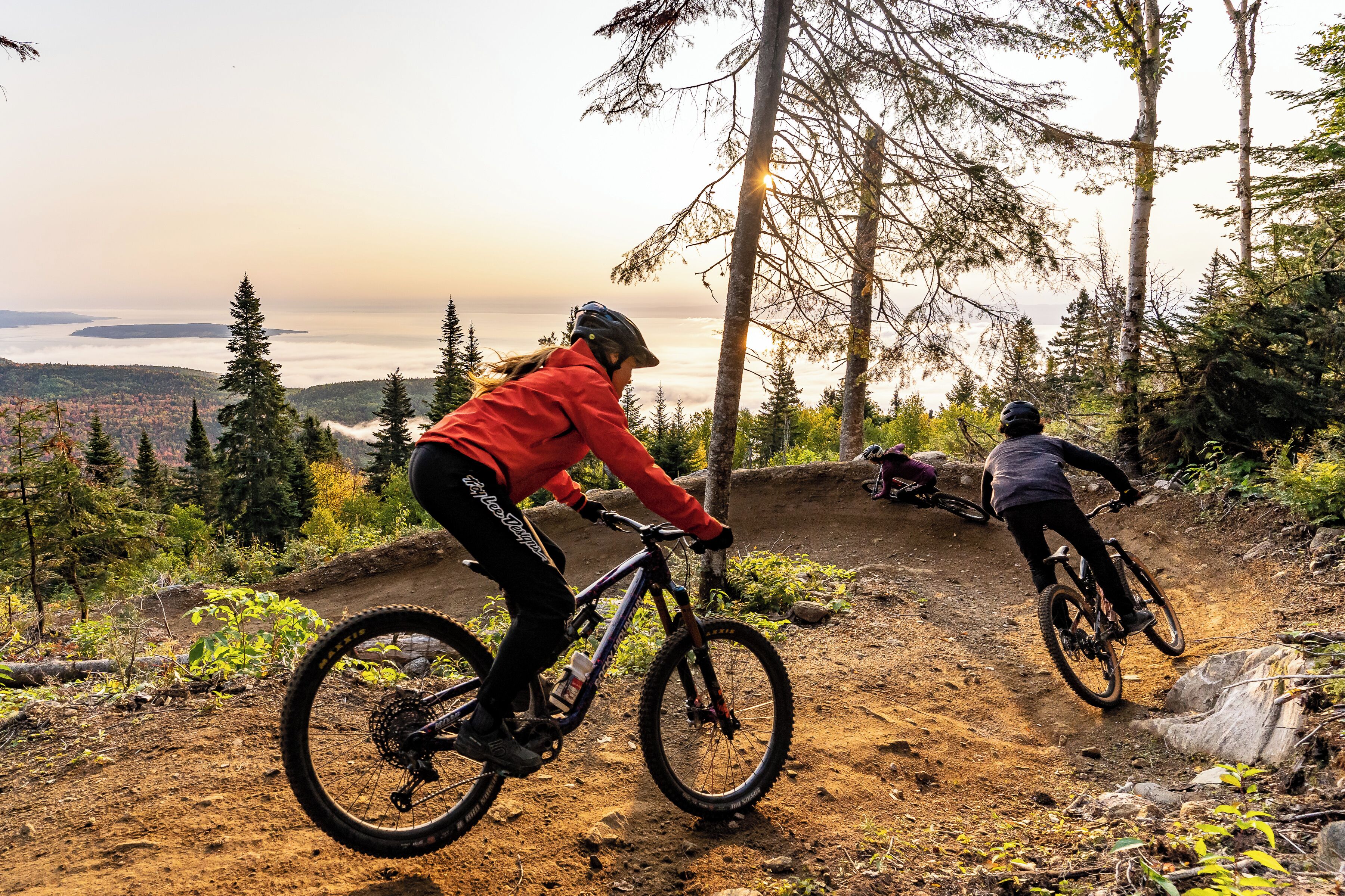 Two mountain bikers on a dirt trail with a scenic view of a forest and a body of water in the background.