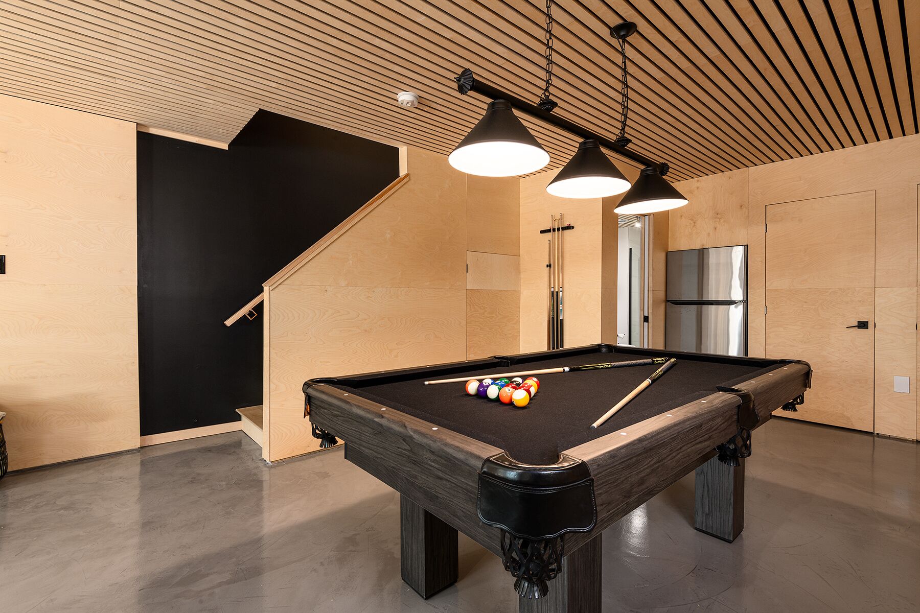 Ein Billardtisch mit Kugeln und Spielstöcken, eine Treppe und ein Kühlschrank in einem Raum mit Holzwänden und -decke.