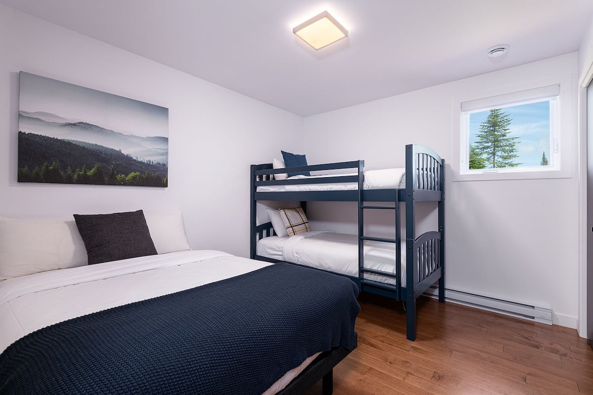 A bedroom with a bunk bed, a single bed, a large landscape painting, and a window with a view of a tree.