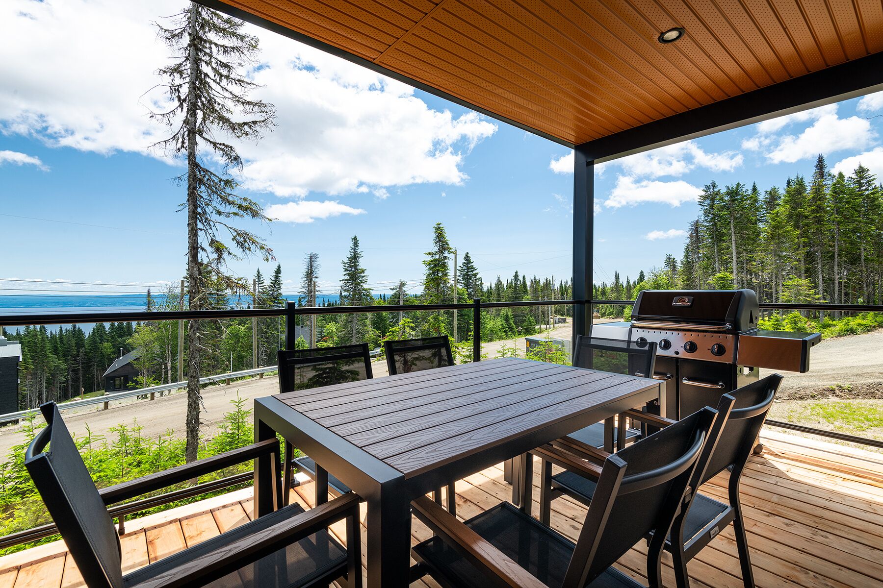 An outdoor deck with a wooden table and chairs, a grill, and a scenic view of a forest and a body of water.