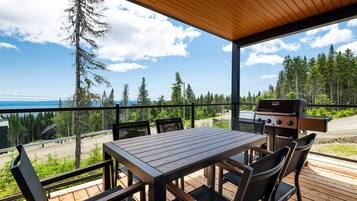 Een terras buiten met een houten tafel en stoelen, een barbecue en een prachtig uitzicht op een bos en een waterlichaam.