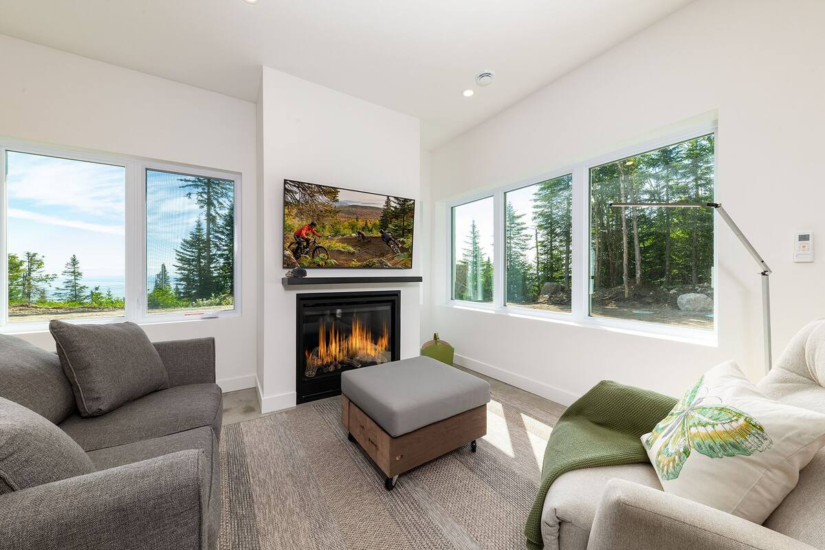 A modern living room with a fireplace, large windows, and a view of a forest.