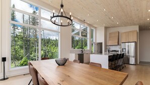 A spacious kitchen with large windows, a wooden dining table, and a modern chandelier. - Les Chalets Forêt (Petite-Riviere-Saint-Francois)