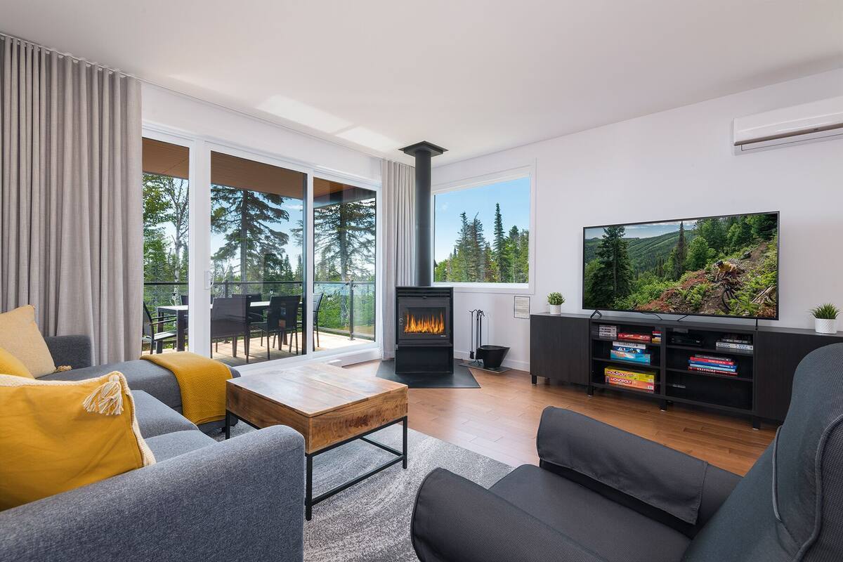 A modern living room with a fireplace, a television, and a balcony with a view of trees.