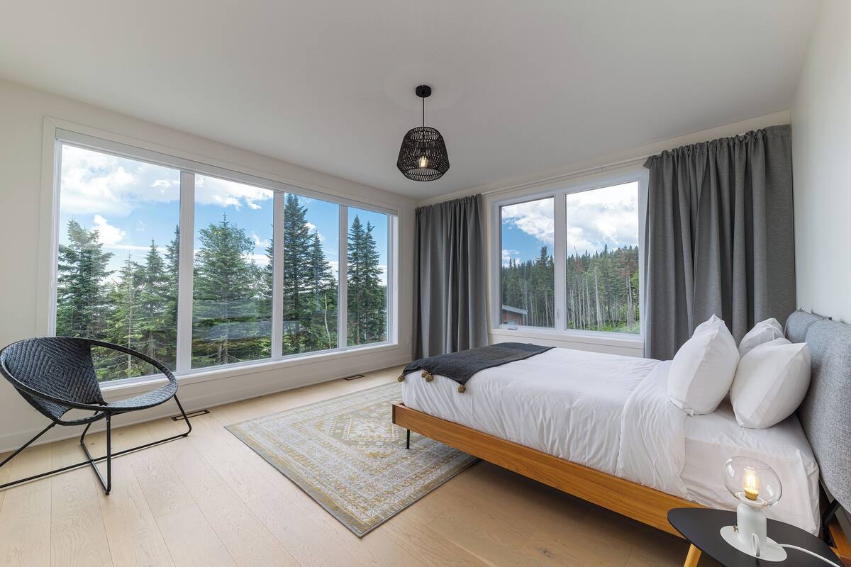 A bedroom with a large window, a bed, a chair, and a bedside table with a lamp.