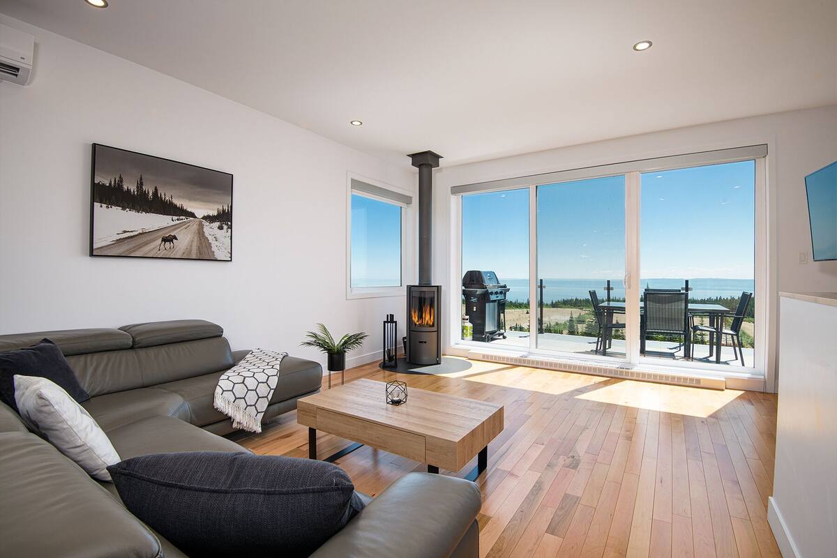 A modern living room with a sofa, coffee table, and a fireplace. There is a large window with a view of the outdoors.
