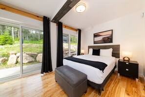 A modern bedroom with a large bed, a bench, a nightstand, and a view of a forested area through large windows. - Les Chalets Forêt (Petite-Riviere-Saint-Francois)