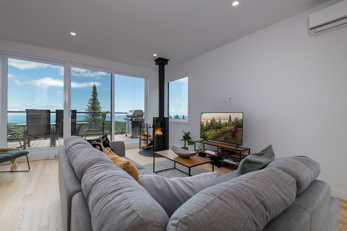A modern living room with a large grey sofa, a fireplace, a television, and a balcony with a view of the ocean.