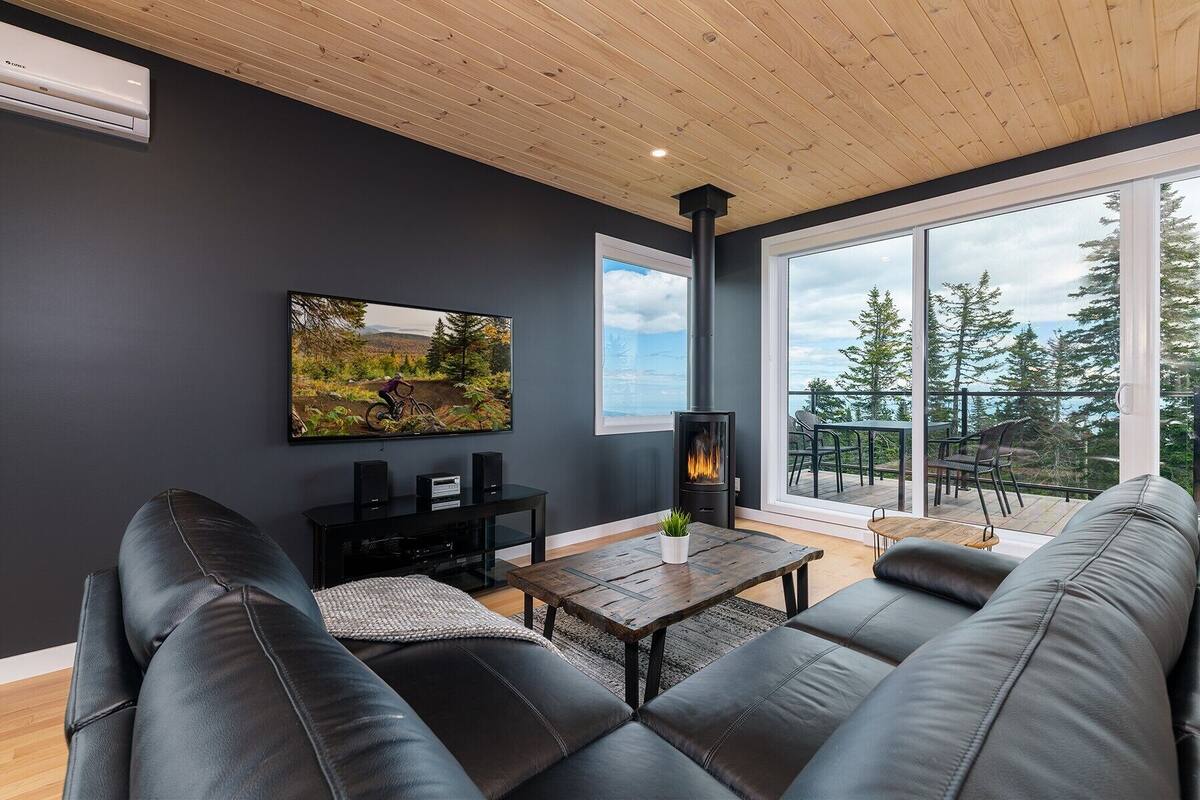 A modern living room with a black leather sofa, a wooden coffee table, a mounted TV, and a wood-burning stove.